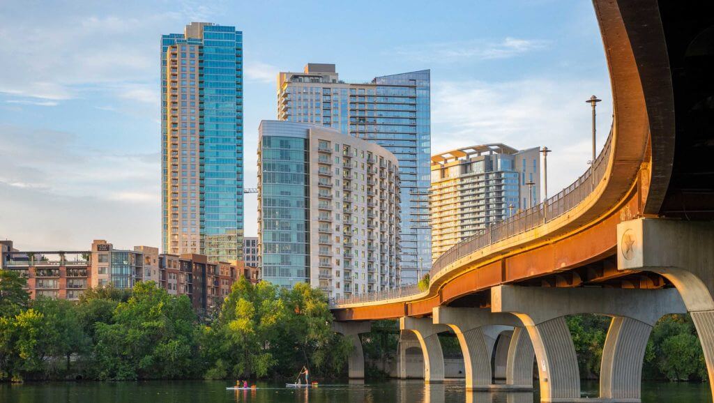 Highrise buildings look over Austin, Texas riverfront and family paddleboarding under a large bridge