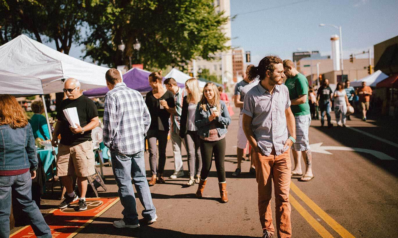 Residents walk around an outdoor market with white and purple tents set up.
