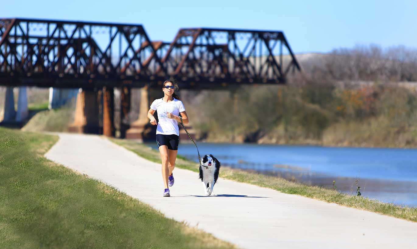 A woman runs alongside her dog on a sidewalk next to a river at Trinity Trails in Fort Worth, Texas.