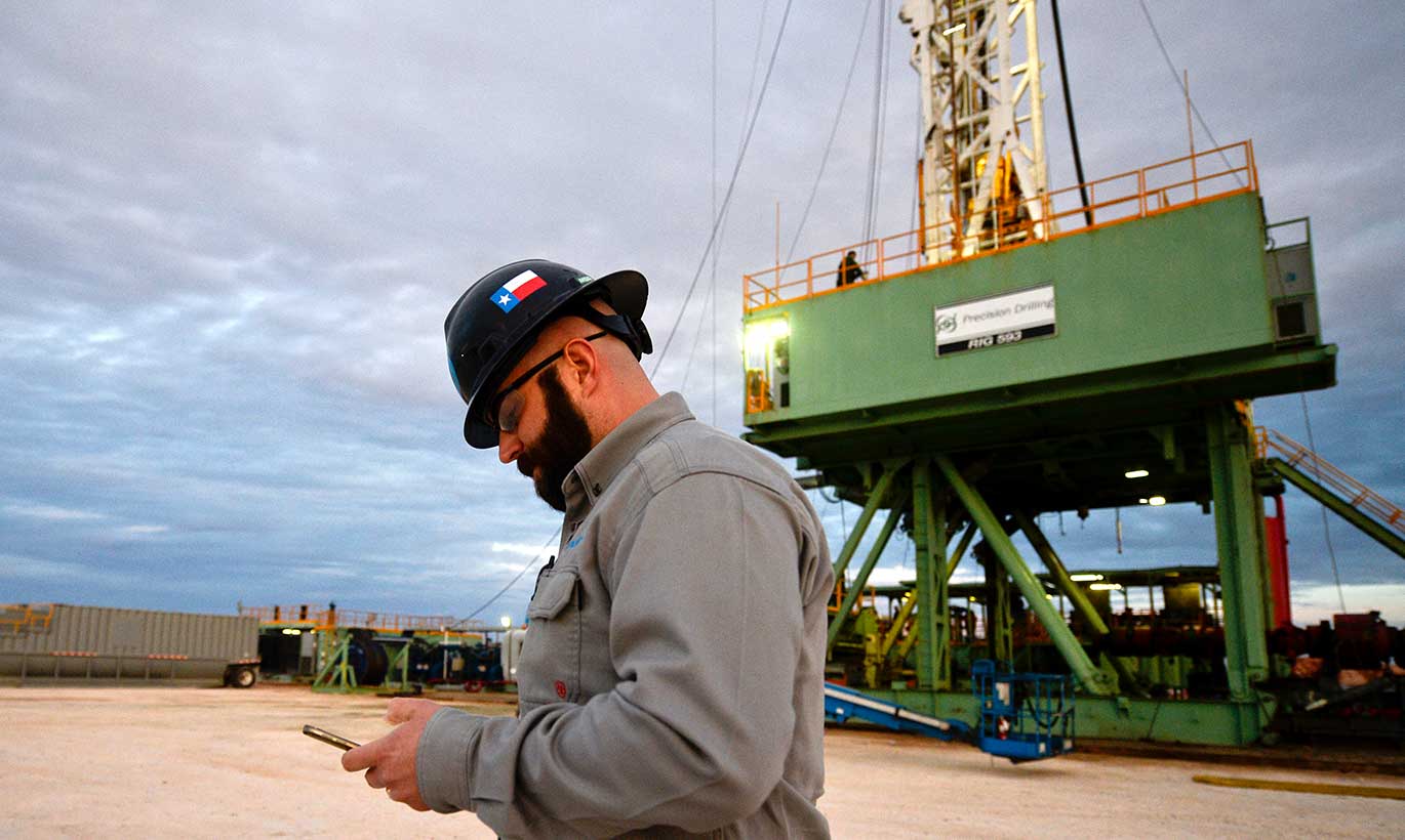 A man wearing a hard hat looks down at his phone on a Precision Drilling work site