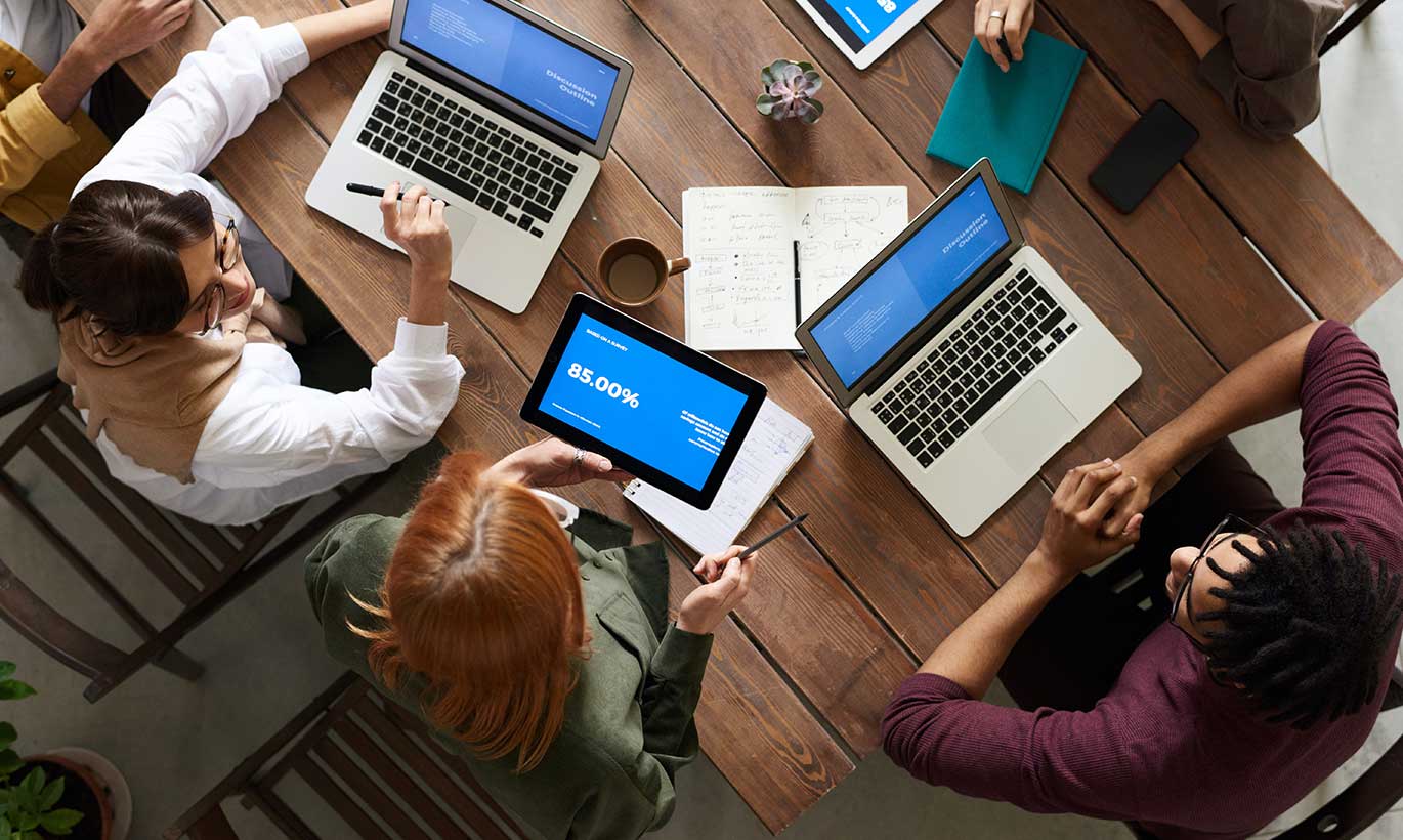 Coworkers sit at a large wood table and work with laptops and digital tablets during a meeting.