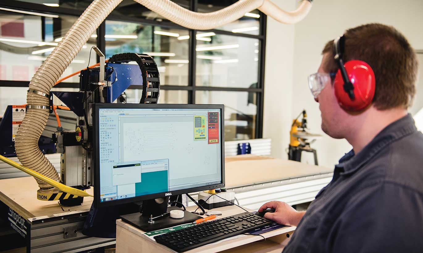 A man wearing protective ear equipment works on a computer in a manufacturing facility.