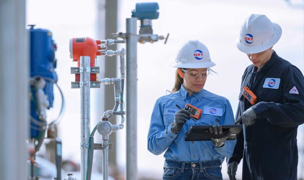 Two Occidental workers work out in the field with a tablet next to industrial equipment